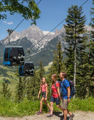 Family hiking on forest trail with gondolas in background and mountain panorama | © Roland Haschka