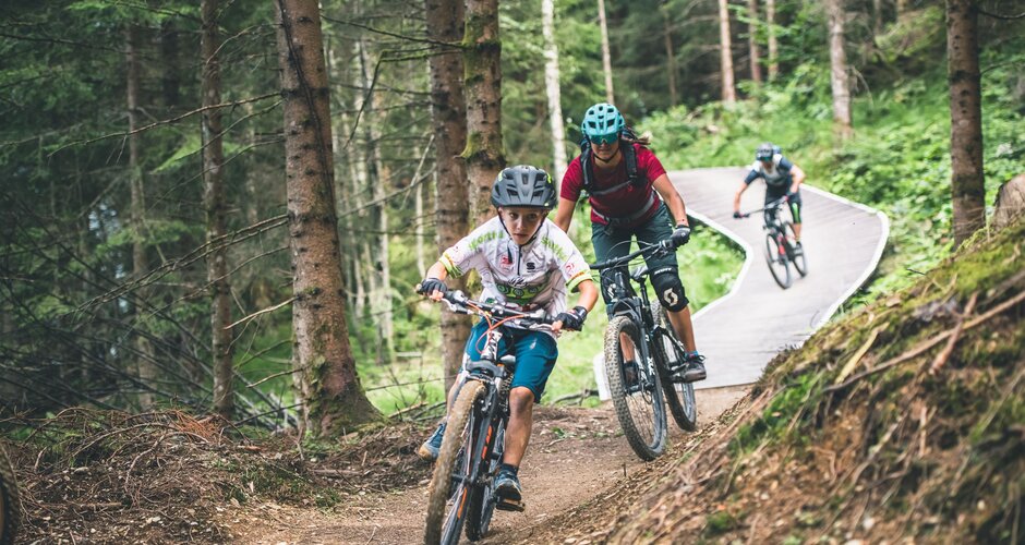 Mountain bikers ride narrow flow trail through forest with wooden bridges and bends