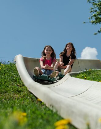 Girl and woman ride down the Biberg summer toboggan track, smiling in the sun, with flowers and blue sky all around. | © Hochkönig / Roland Haschka