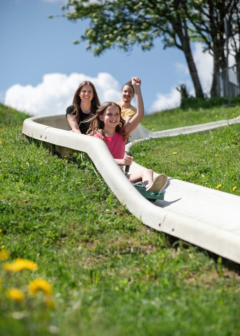Smiling girl rides the summer toboggan run down a grassy slope with her family, wildflowers visible in the foreground. | © Hochkönig / Roland Haschka
