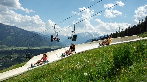 Three people ride the Biberg summer toboggan run, lush green hills and a chairlift above, mountains in the background. | © Hochkönig / Roland Haschka
