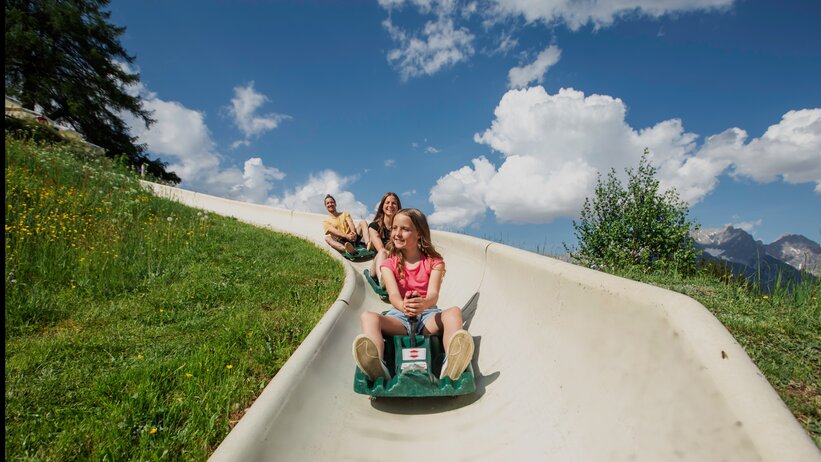 Girl rides the Biberg summer toboggan with two adults behind her, green slope and blue sky in a scenic alpine landscape. | © Hochkönig / Roland Haschka