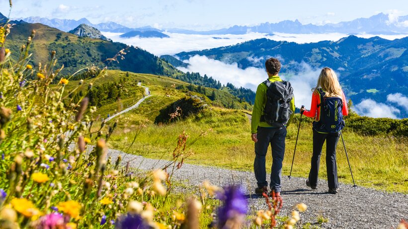 Zwei Wanderer mit Rucksäcken auf Almweg, bunte Blumen im Vordergrund, Berge und Wolkenmeer im Hintergrund | © Dorfgasteiner Bergbahnen AG