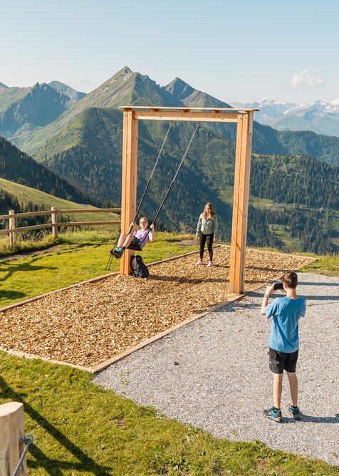 Jugendliche schaukeln vor Bergpanorama, andere machen Pause oder fotografieren am Gipfel | © Dorfgasteiner Bergbahnen AG