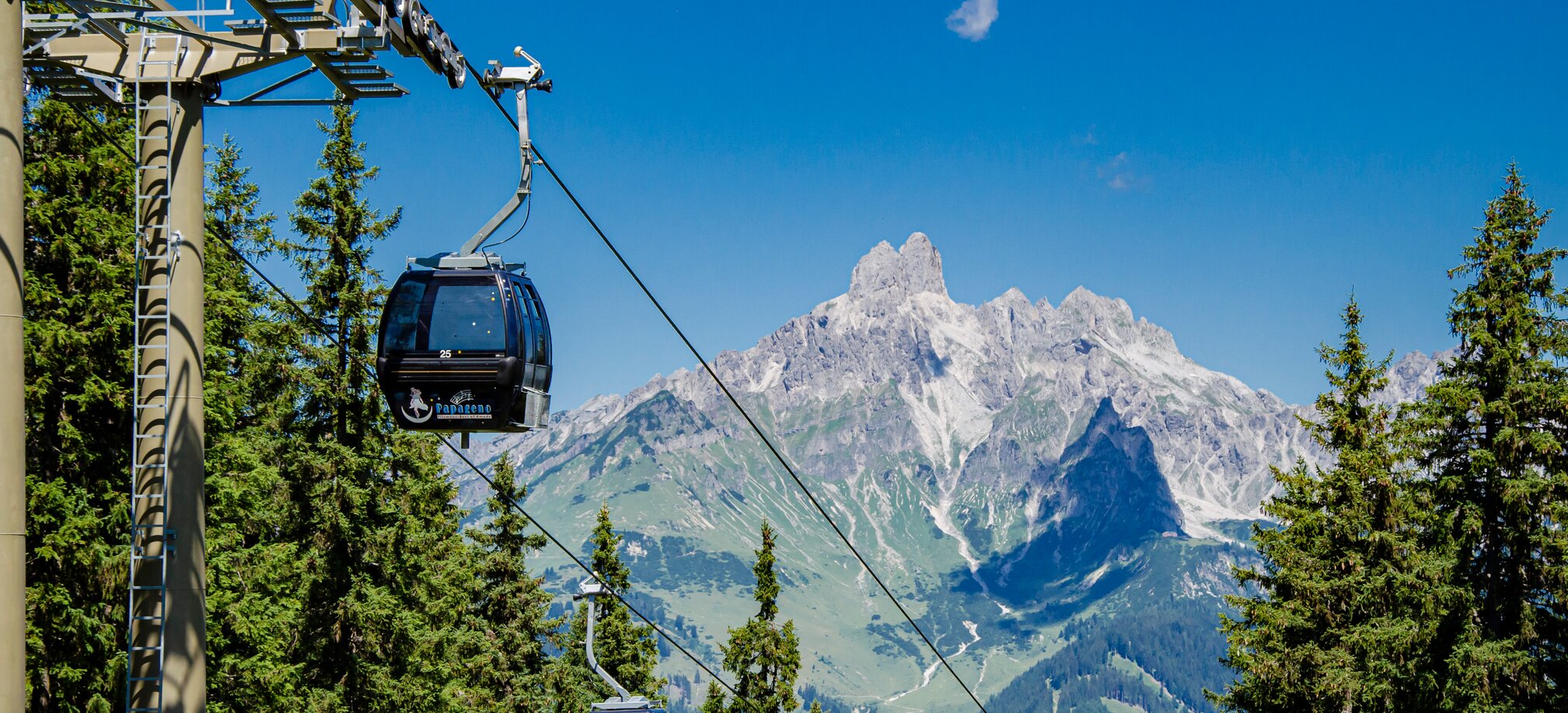 Papagenobahn cable cars schweben über Nadelwald in Filzmoos, dahinter ragt ein markantes Felsmassiv auf. | © filzmoos.ski