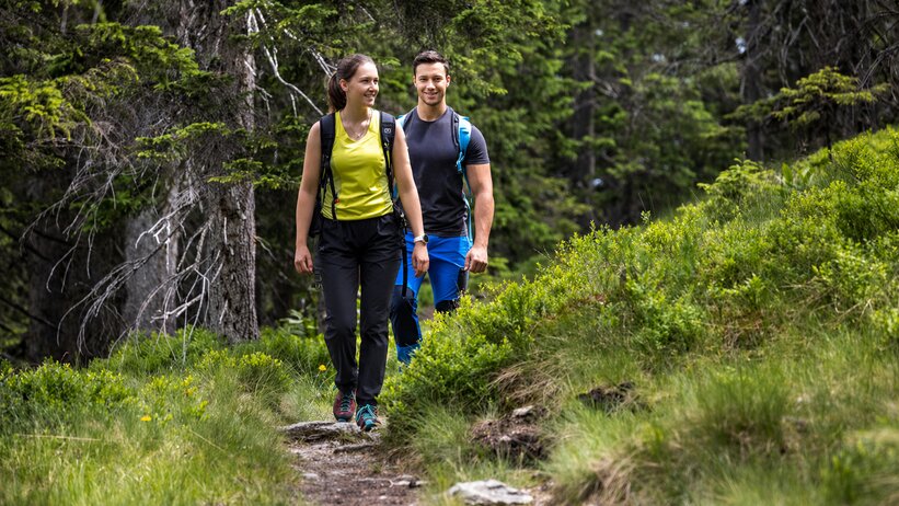 Zwei Wanderer gehen auf schmalem Waldweg durch grüne Wiesen und dichte Nadelbäume in Filzmoos im Sommer. | © filzmoos.ski_Christian Fischbacher