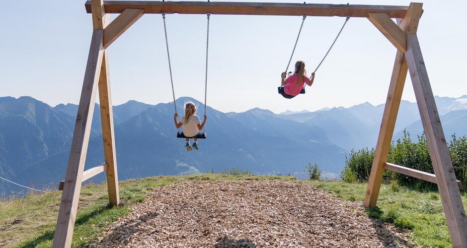 Zwei Kinder schaukeln auf Holzschaukel vor Bergpanorama mit Fernblick über das sonnige Gasteinertal | © Gasteiner Bergbahnen AG