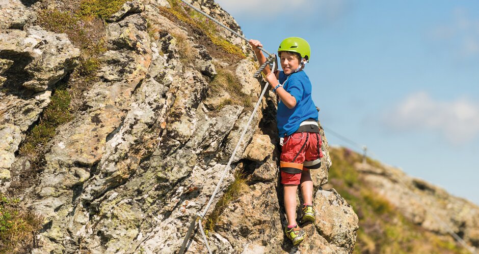 Boy with helmet and harness climbing on secured rock face in sunny alpine landscape | © Gasteiner Bergbahnen AG