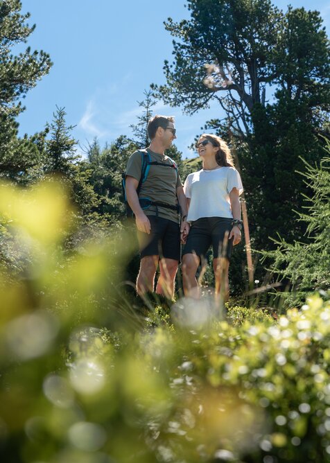 Lachendes Paar wandert am Graukogel durch Wald im Sonnenschein, umgeben von grüner Bergnatur | © Skigastein, seven frames