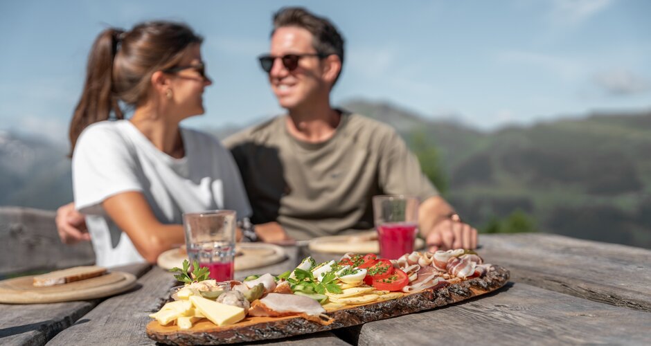 Paar genießt Brettljause mit Käse, Speck und Saft auf Holztisch vor Bergpanorama | © Skigastein, seven frames