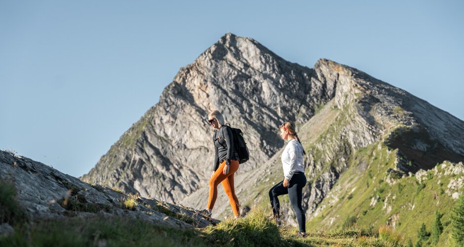 Zwei Frauen wandern am Graukogel bergauf, im Hintergrund ein markanter, felsiger Berggipfel bei klarem Himmel | © Skigastein, seven frames