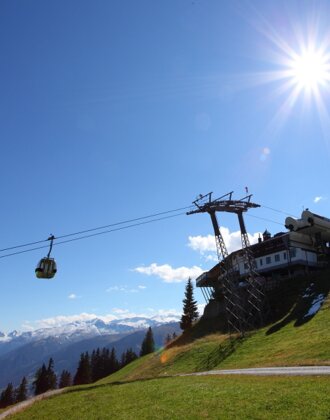 Cable car ascends to mountain station on sunny day, with green meadow, snowy mountain backdrop and clear blue sky. | © Großarler Bergbahnen