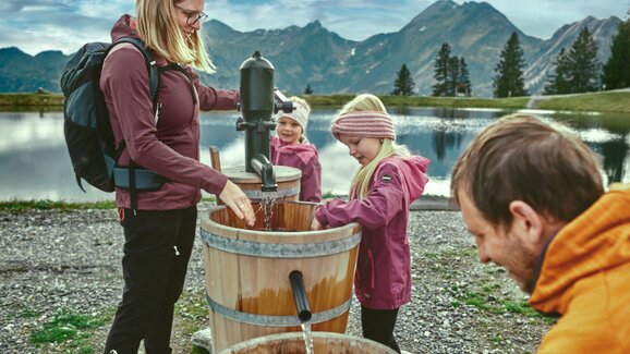 Family with kids pumping water from wooden barrels by a lake in Großarl, mountains and clouds reflecting in the background. | © Großarler Bergbahnen