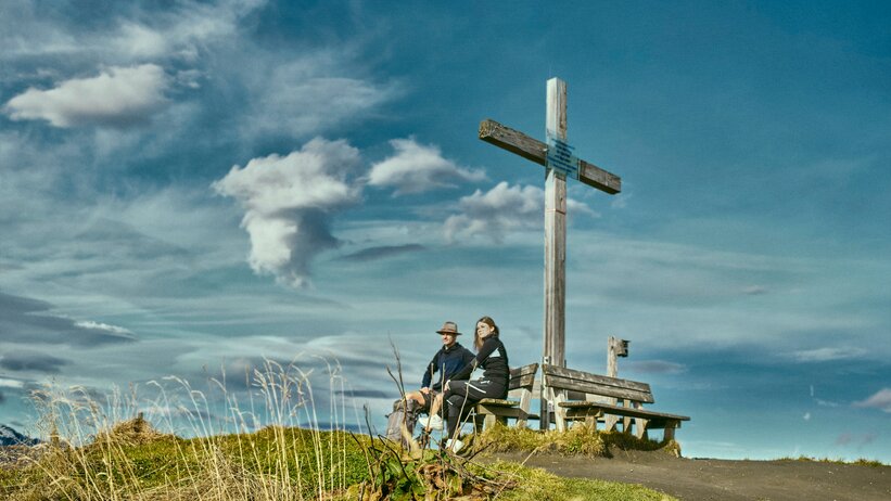 Two people rest on a bench by the summit cross in Großarl, enjoying the view beneath a dramatic, partly cloudy sky. | © Großarler Bergbahnen