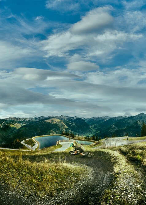 Mountain biker on winding trail overlooking reservoir, forested slopes and a sky full of dramatic clouds. | © Großarler Bergbahnen