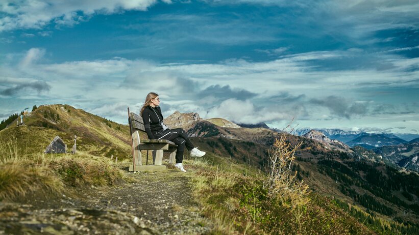 Woman sits on a bench along a mountain path in Großarl, overlooking autumn hills and a dramatic cloudy sky. | © Großarler Bergbahnen