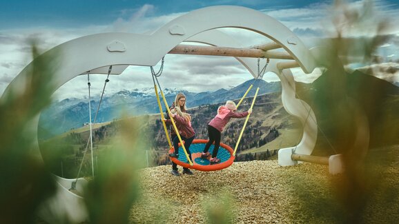 Two kids play on a large nest swing with a mountain backdrop in Großarl, wood chips on the ground, clouds in the sky. | © Großarler Bergbahnen