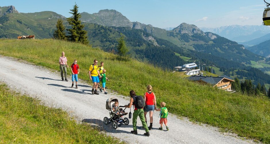 Families hiking with children and stroller on a wide mountain path, cable car in background, green hills and distant view. | © Großarler Bergbahnen