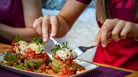 Close-up of hands cutting spinach dumplings with tomato sauce and cheese on a plate in a rustic mountain hut. | © Hauser Kaibling