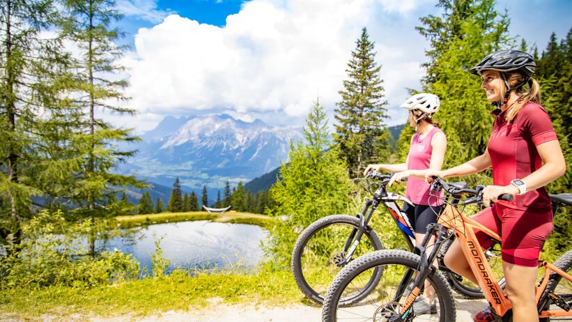 Two women with mountain bikes enjoy the view at a mountain lake | © Hauser Kaibling