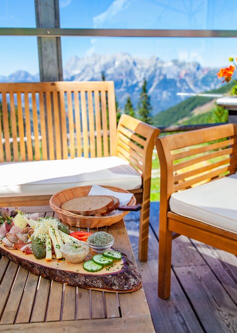 Wooden table with snack platter, bread basket and menu on sunny terrace with mountain view and blooming geraniums. | © René Eduard Perhab