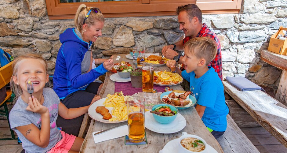 Family sits on wooden benches by stone wall enjoying schnitzel, fries and local dishes during an alpine hut visit. | © René Eduard Perhab