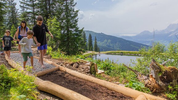 Family with kids walking barefoot on a trail of gravel and soil at Hauser Kaibling, with mountain lake and Alps in the background. | © René Eduard Perhab
