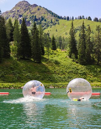 Two kids inside transparent zorbing balls on a lake with green mountains in the background. | © Hauser Kaibling