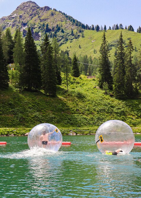 Two kids inside transparent zorbing balls on a lake with green mountains in the background. | © Hauser Kaibling