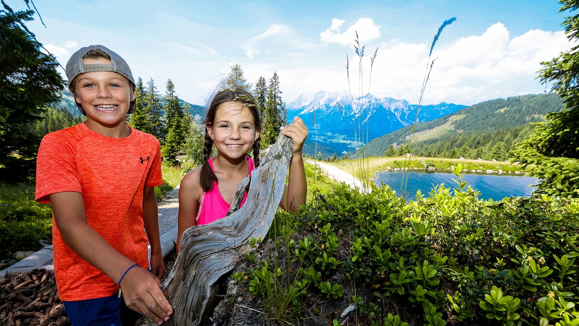 Two smiling kids by a tree stump at Schafsinn pond with mountain views in summer. | © Stefan Pajman