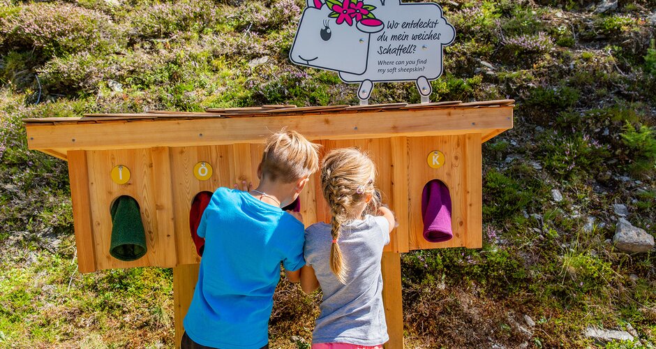 Two kids feel into a wooden tactile station with fabrics; a sheep sign presents a riddle on the themed mountain trail. | © Hauser Kaibling