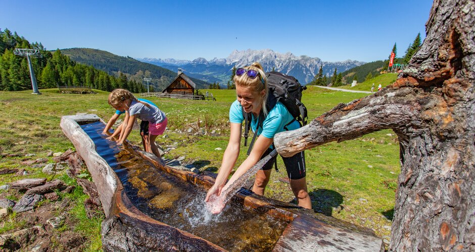 Woman and girl refresh at a wooden water trough while hiking, with alpine landscape and hut in the background. | © Hauser Kaibling