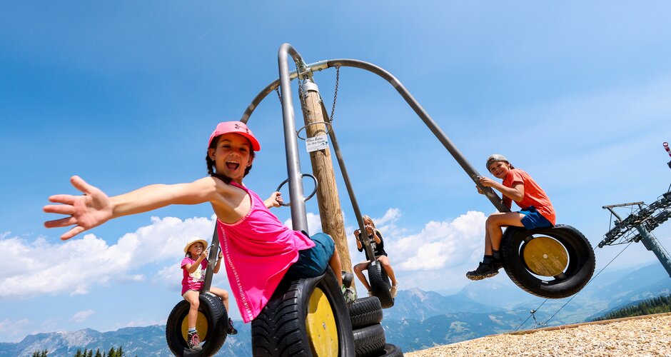Kids swinging on a tire carousel with mountain view, laughing and stretching arms happily toward the camera. | © Stefan Pajman