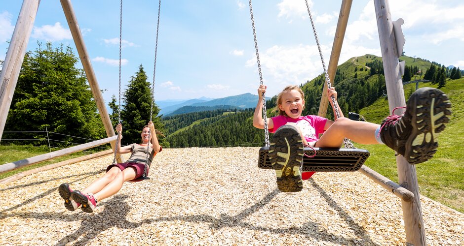 Smiling child and woman on wooden swings with view of green mountains, forest landscape and sunny blue sky in the background. | © Stefan Pajman