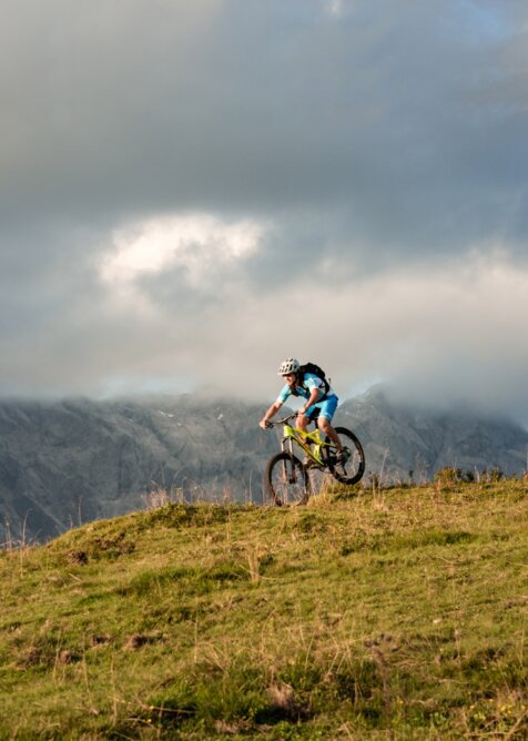 Mountainbiker fährt über Almwiese vor wolkenverhangenem Hochkönigmassiv im abendlichen Licht | © Christian Schartner