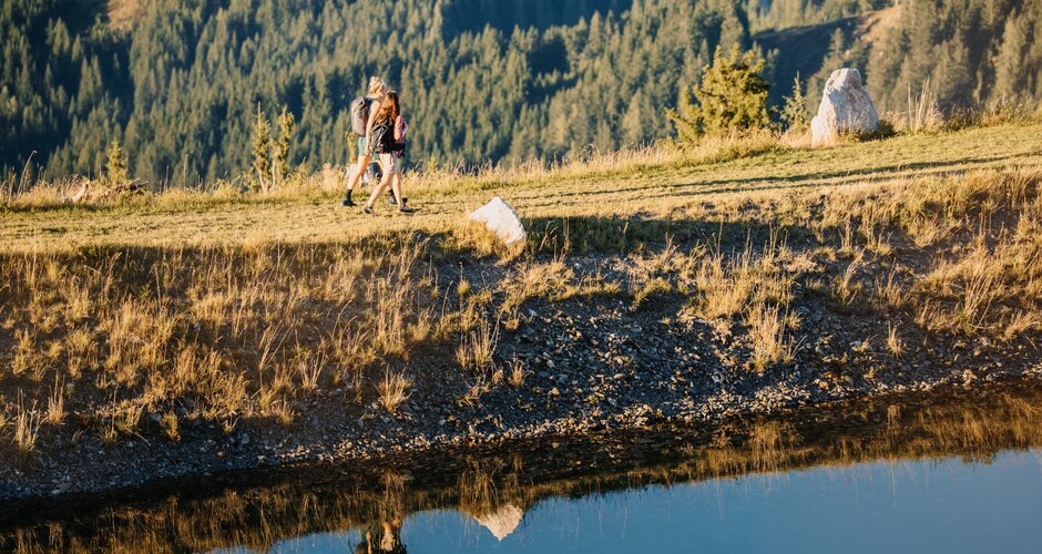 Two hikers walk along the edge of a small pond near Bürglalmbahn in Dienten across a grassy slope. | © Hochkönig Bergbahnen