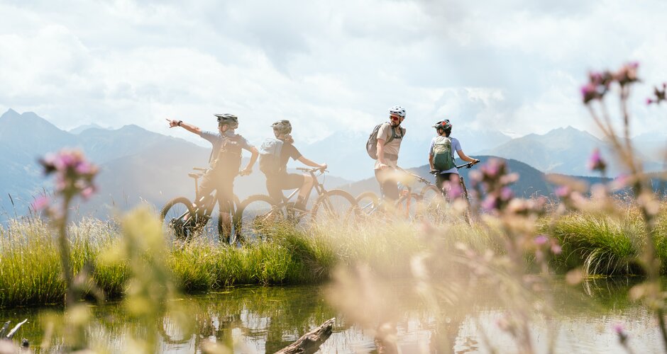 Four mountain bikers stand with their bikes by a small pond with mountains behind near Bürglalmbahn in Dienten | © Hochkönig Bergbahnen