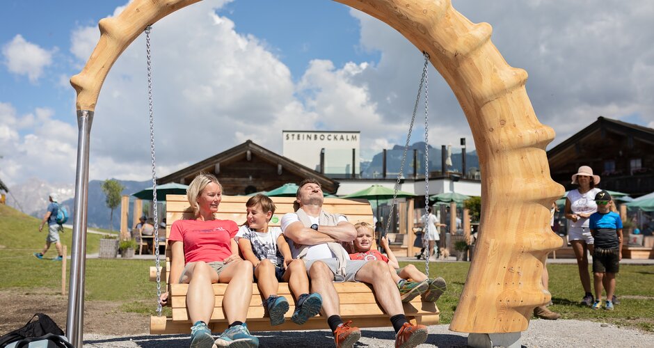 Familie sitzt entspannt auf großer Holzschaukel mit geschnitztem Bogen vor der Steinbockalm im Schaukelpark Hochkönig | © Manu Lochner
