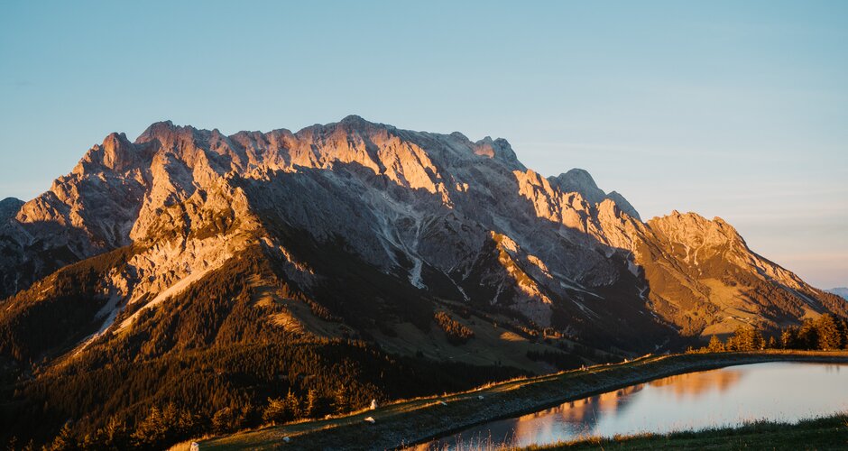 Sunlight illuminates a rocky mountain massif above a reservoir in Dienten beneath a clear evening sky. | © Hochkönig Bergbahnen