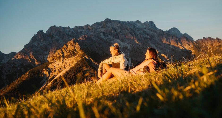 Two female hikers sit on a grassy slope in evening light in Dienten with a rocky mountain massif near Gabühelbahn. | © Hochkönig Bergbahnen