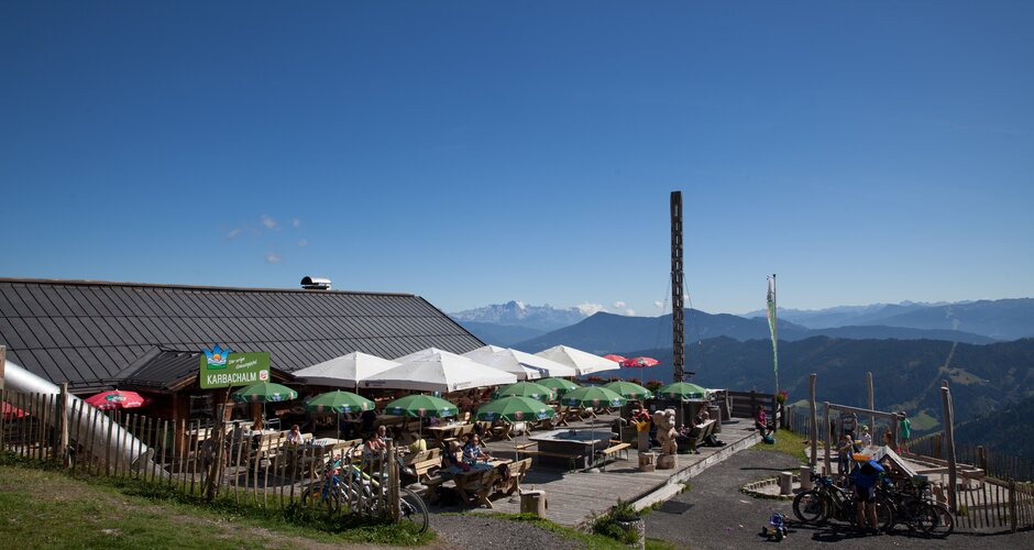 Terrasse der Karbachalm mit Sonnenschirmen, Spielplatz, Ausblick auf die Berge und Gästen an Tischen bei Sonnenschein