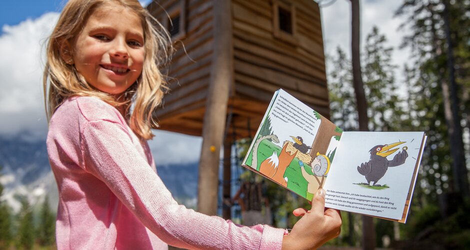 Mädchen zeigt ein aufgeschlagenes Kinderbuch vor Holzhütte im Almspielplatz Hochkönig mit Bäumen und Bergen im Hintergrund