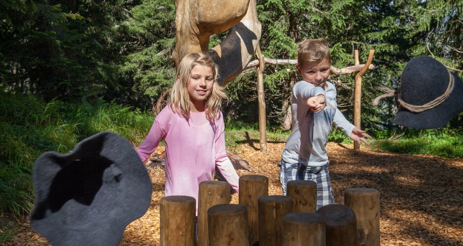 Zwei Kinder spielen ein Hutschnappen-Spiel mit Holzstämmen am Almspielplatz im Wald bei der Karbachalm im Hochkönig