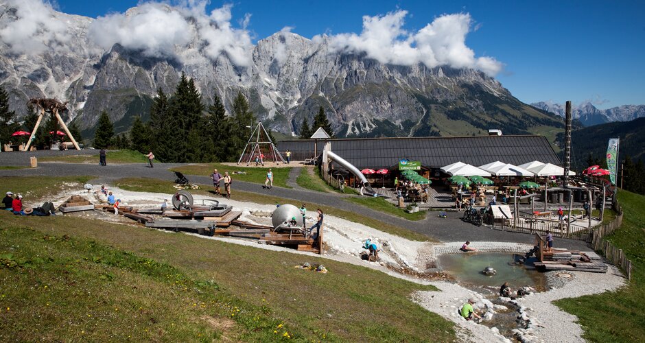 Kinder spielen am Wasserspielplatz bei der Karbachalm, dahinter Terrasse und Hochkönig-Massiv bei Sonnenschein