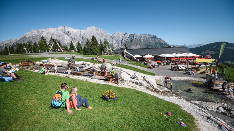 Weitläufiger Almspielplatz in Mühlbach mit Wasserbereich, Terrasse, Gästen und Bergmassiv im Hintergrund. | © Hochkönig Bergbahnen