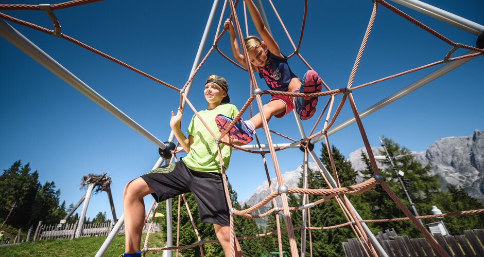 Zwei Kinder klettern im Seilnetzspielplatz unter blauem Himmel mit Alpenpanorama am Hochkönig