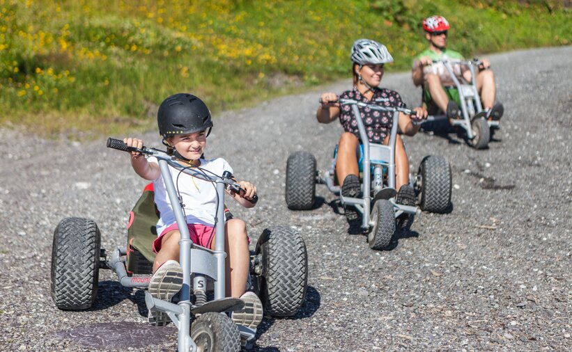 Kind und Erwachsene fahren mit Helmen auf Mountaincarts über eine Schotterstraße in sommerlicher Berglandschaft