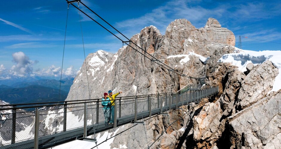 Zwei Personen überqueren die Hängebrücke am Dachstein mit Blick auf schneebedeckte Gipfel und schroffe Felslandschaft bei Sonne. | © Harald Steiner