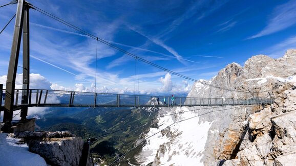 Personen überqueren Hängebrücke am verschneiten Dachstein, mit weitem Blick ins grüne Ennstal unter blauem Himmel. | © Harald Steiner
