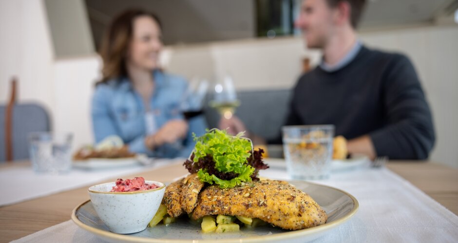 Nahaufnahme eines panierten Fischfilets mit Salat und Beilage, im Hintergrund unscharf lachendes Paar mit Weingläsern. | © Harald Steiner